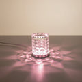 A lit cylindrical table lamp with a faceted textured glass shade and a silver base, casting a pink glow on a glass table next to stacked vintage books.